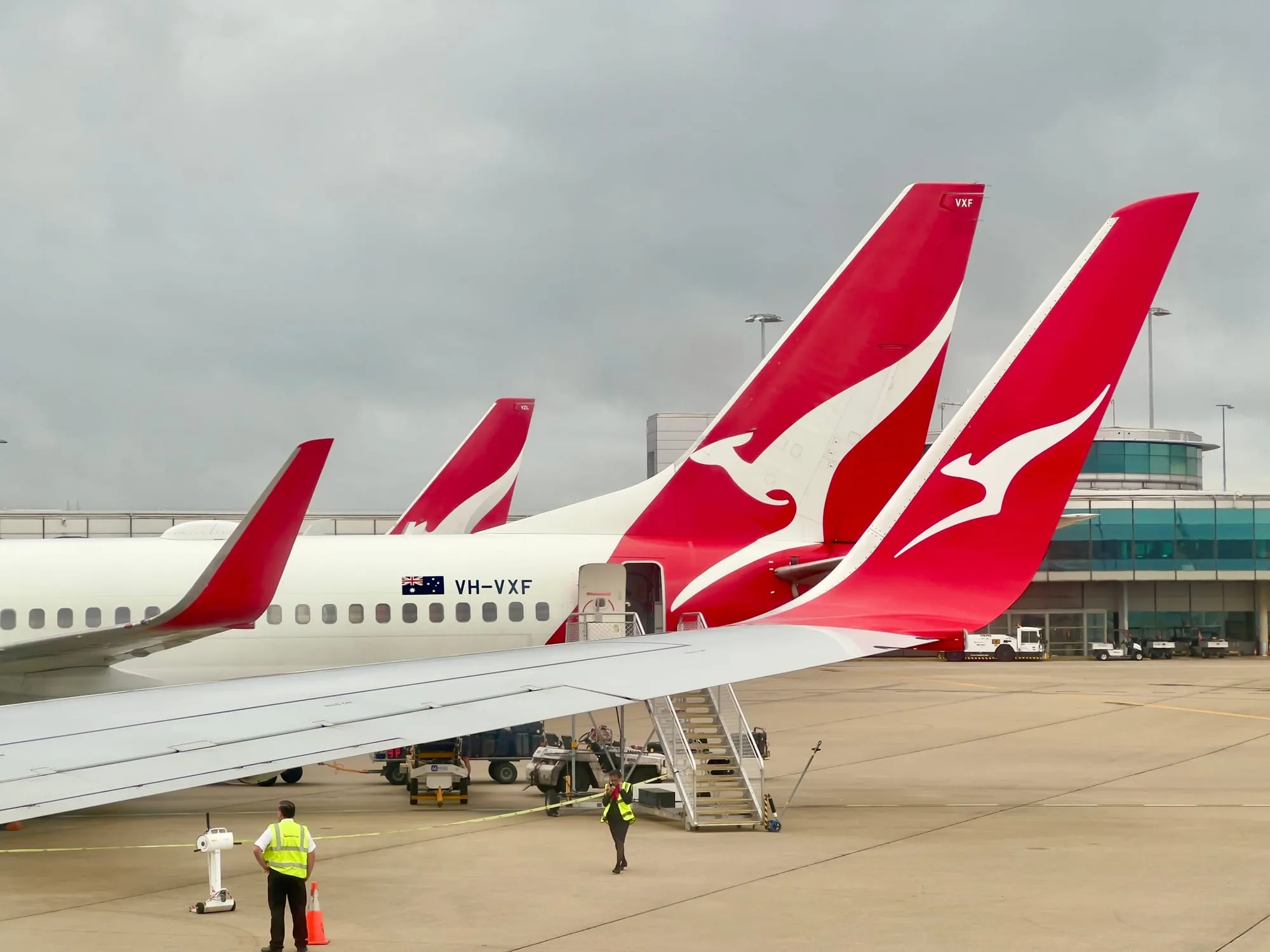 Four Qantas Boeing 737 tails lined up at gates under an overcast sky, ground crew in high-visibility vests working beneath the nearest wing.