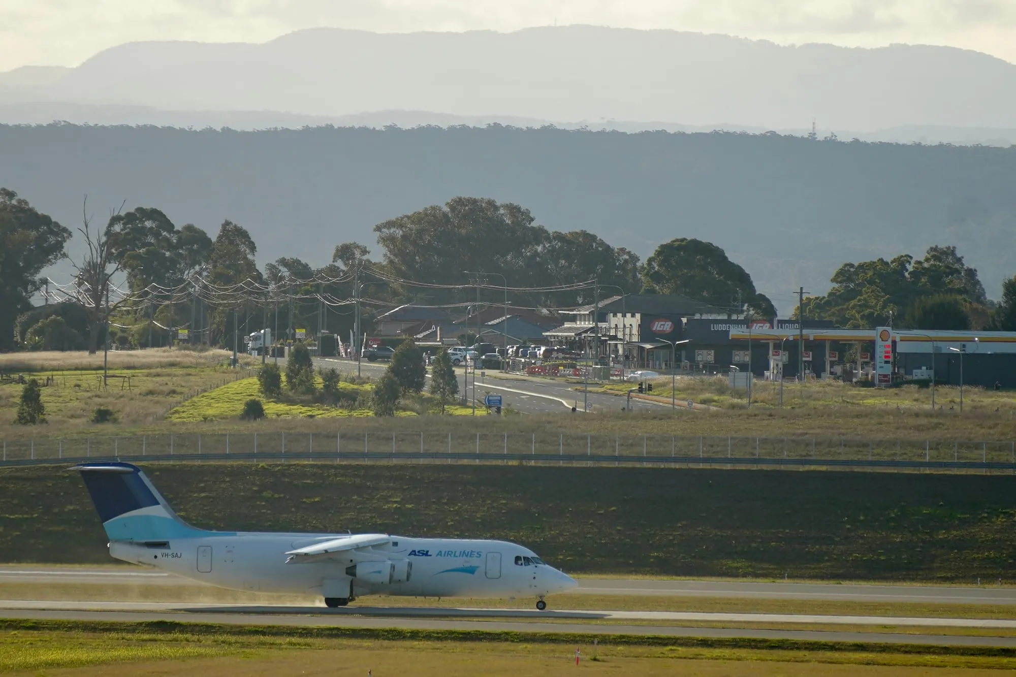 A four-engined BAe 146 freighter taxiing along a runway, with the shopfronts of a small village — a general store, a liquor shop and a service station — directly behind the airport fence, and the Blue Mountains beyond.