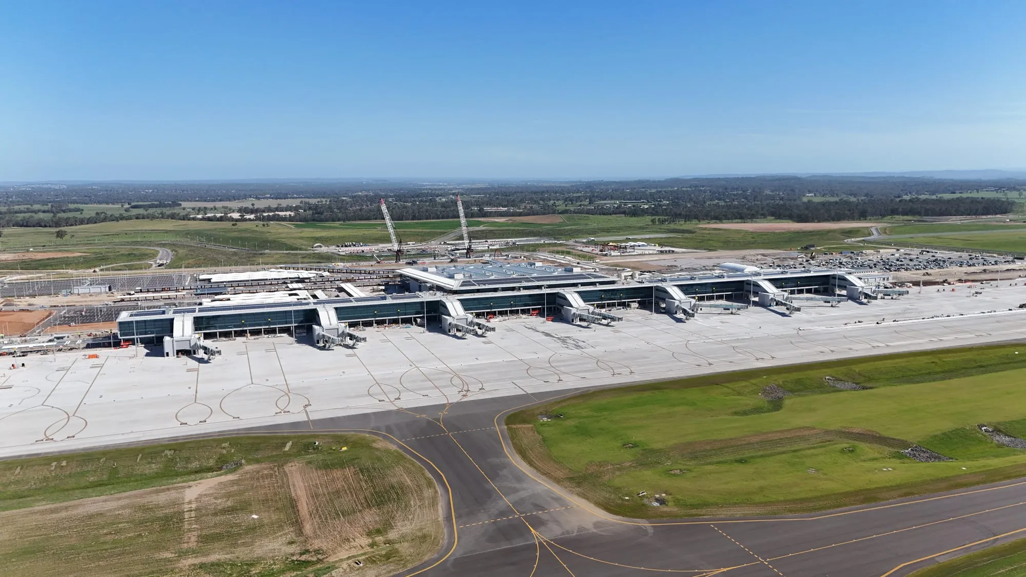 Aerial view of Western Sydney Airport's terminal: a low linear building with a row of aerobridges along a newly poured concrete apron, construction cranes still standing behind it and farmland to the horizon.